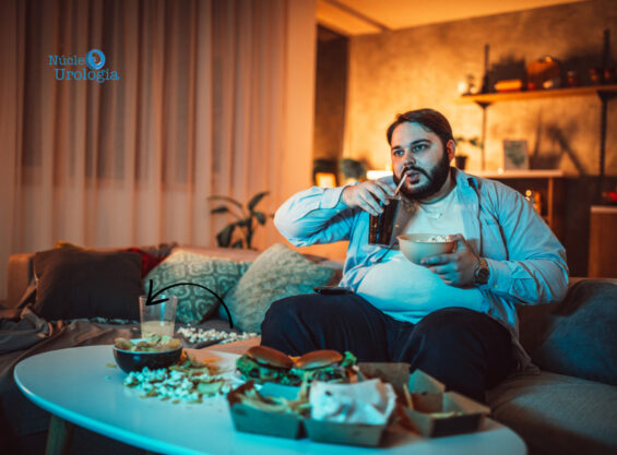 Alimentação pode causar cálculos renais. Homem obeso sentado no sofá e comendo fast food enquanto assite televisão.